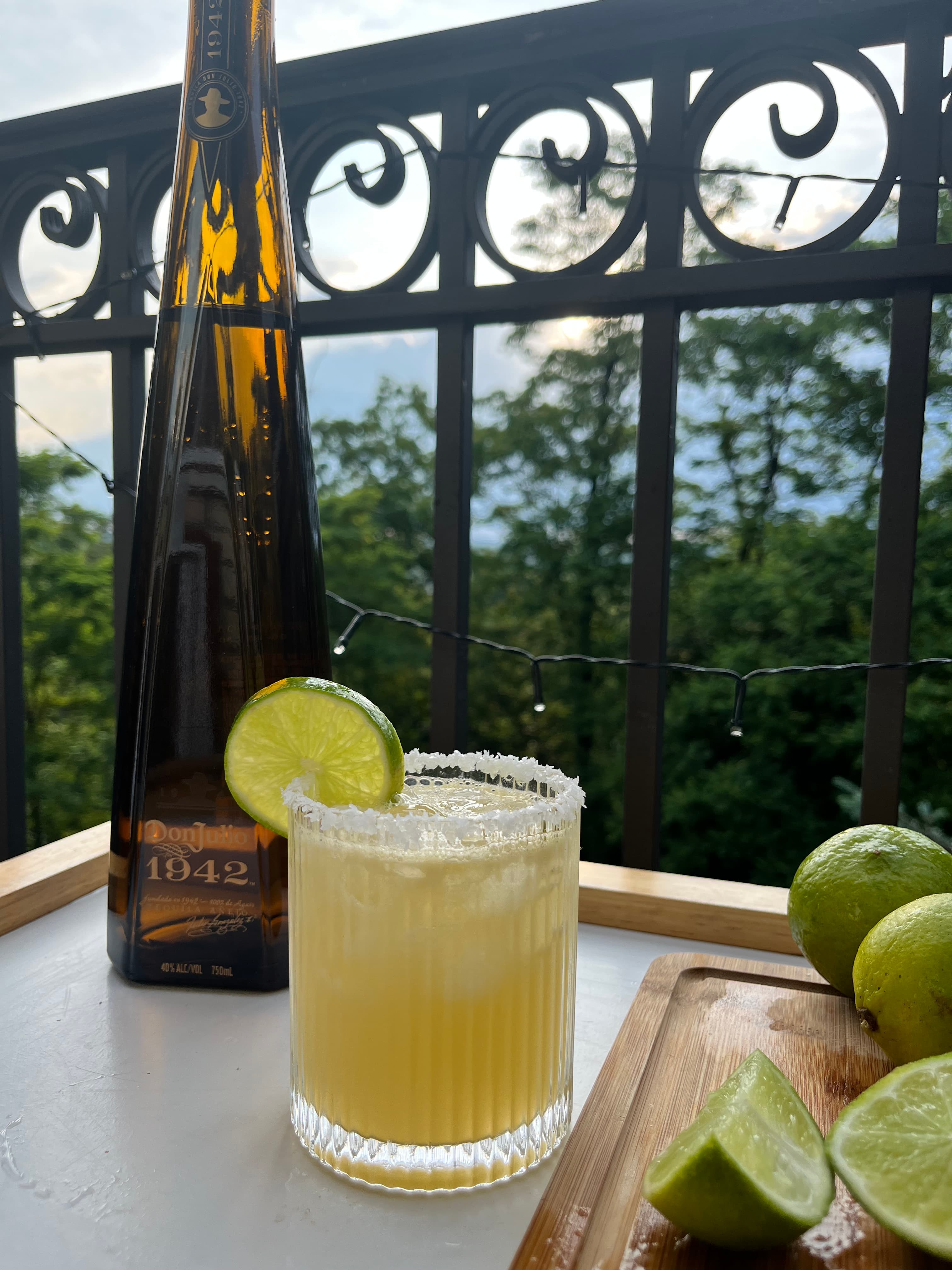A pale yellow cocktail with a salted rim and lime garnish in a textured glass