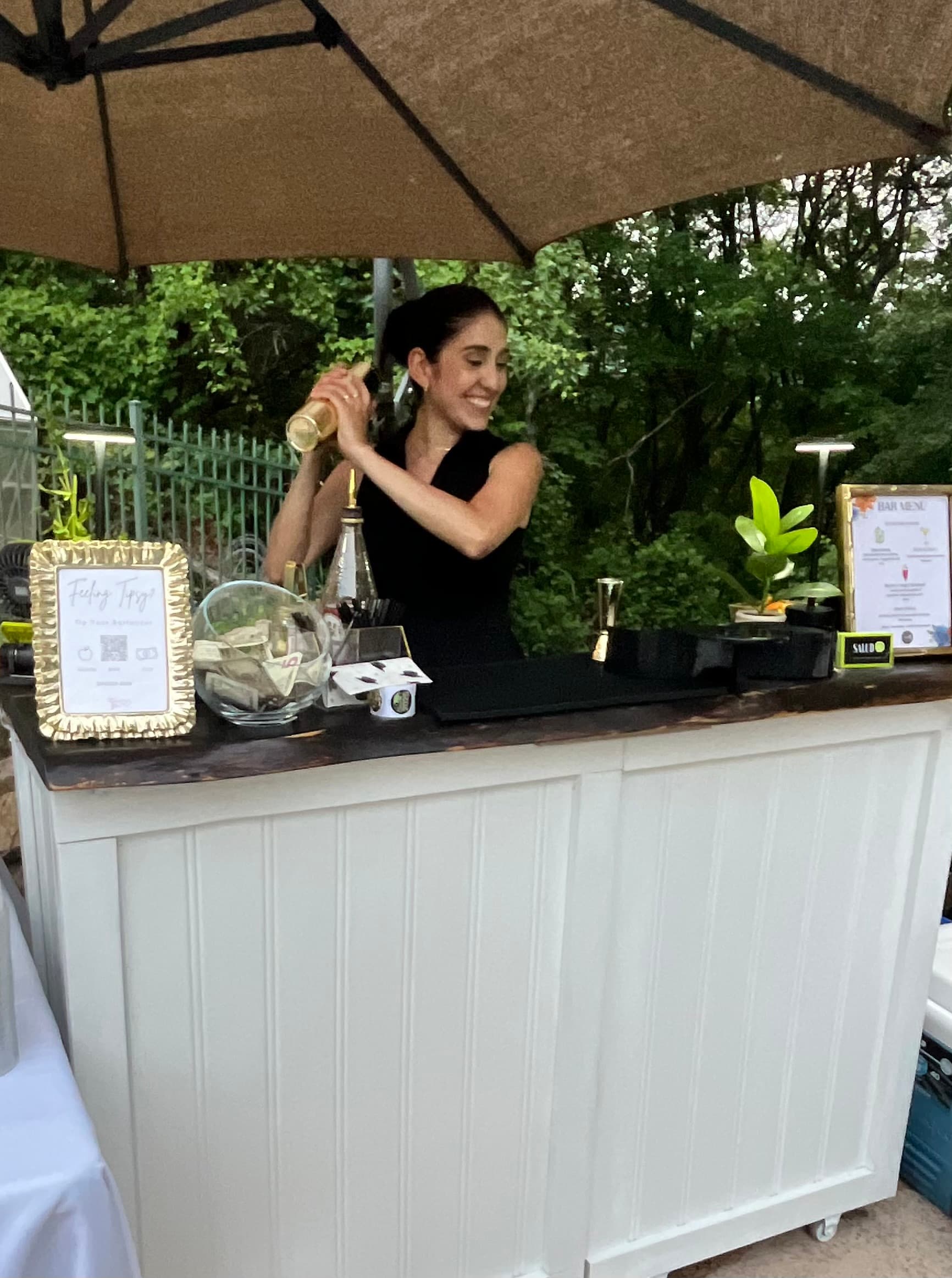 A smiling bartender shaking a fresh cocktail at an outdoor bar.
