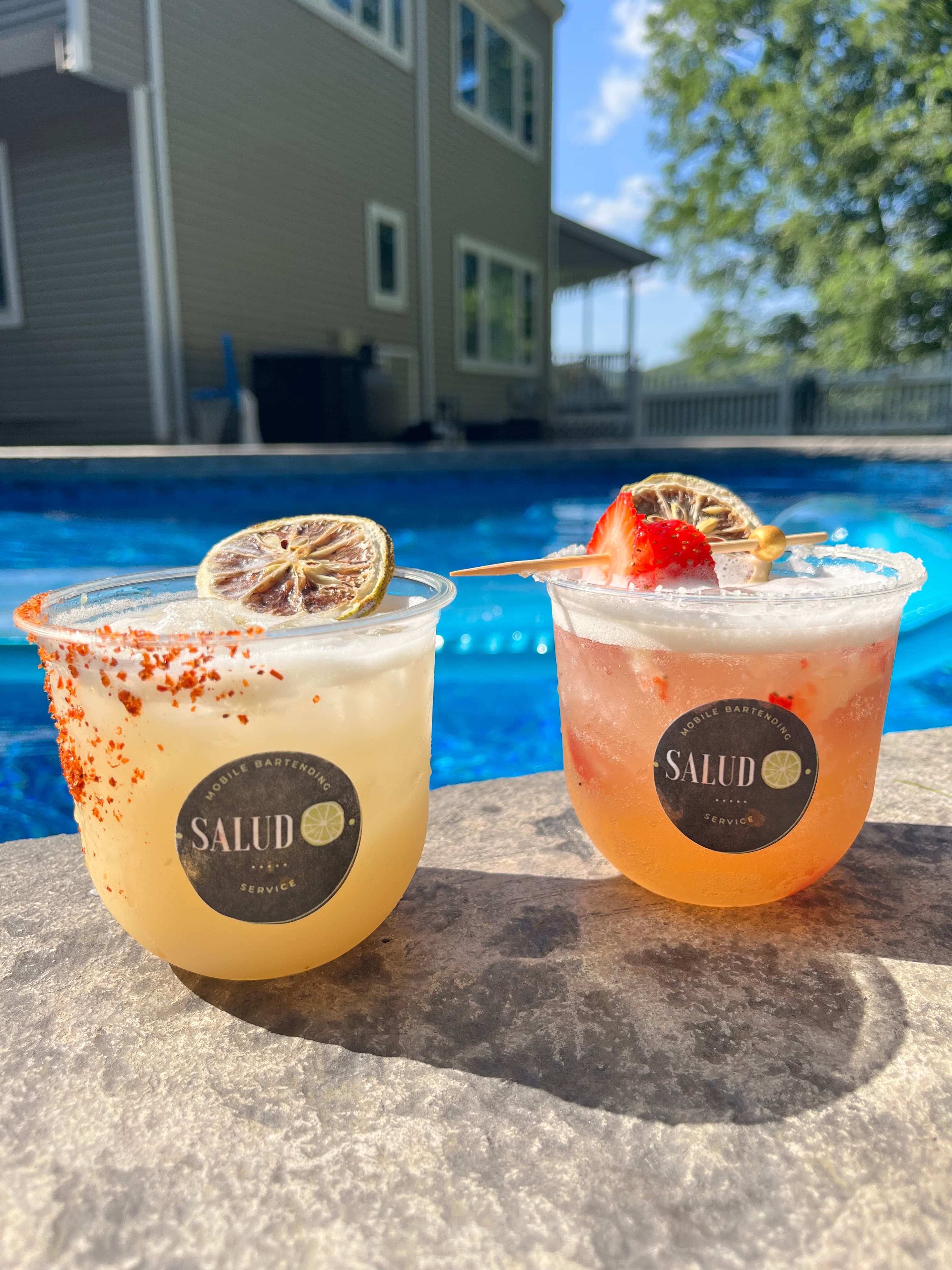 Two handcrafted cocktails with fruit garnishes and salted rims by a pool.